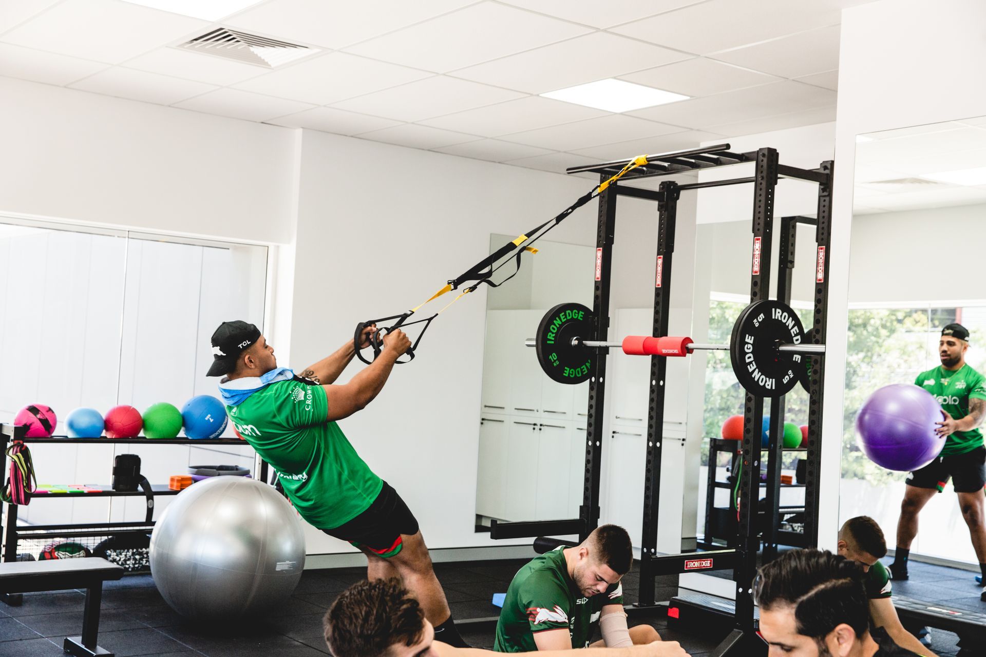 Man in green shirt using suspension straps in gym, another holding exercise ball.