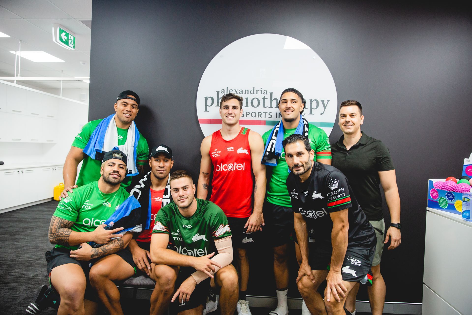 Group of men posing for a photo in a physiotherapy clinic, wearing sports jerseys and towels, smiling.
