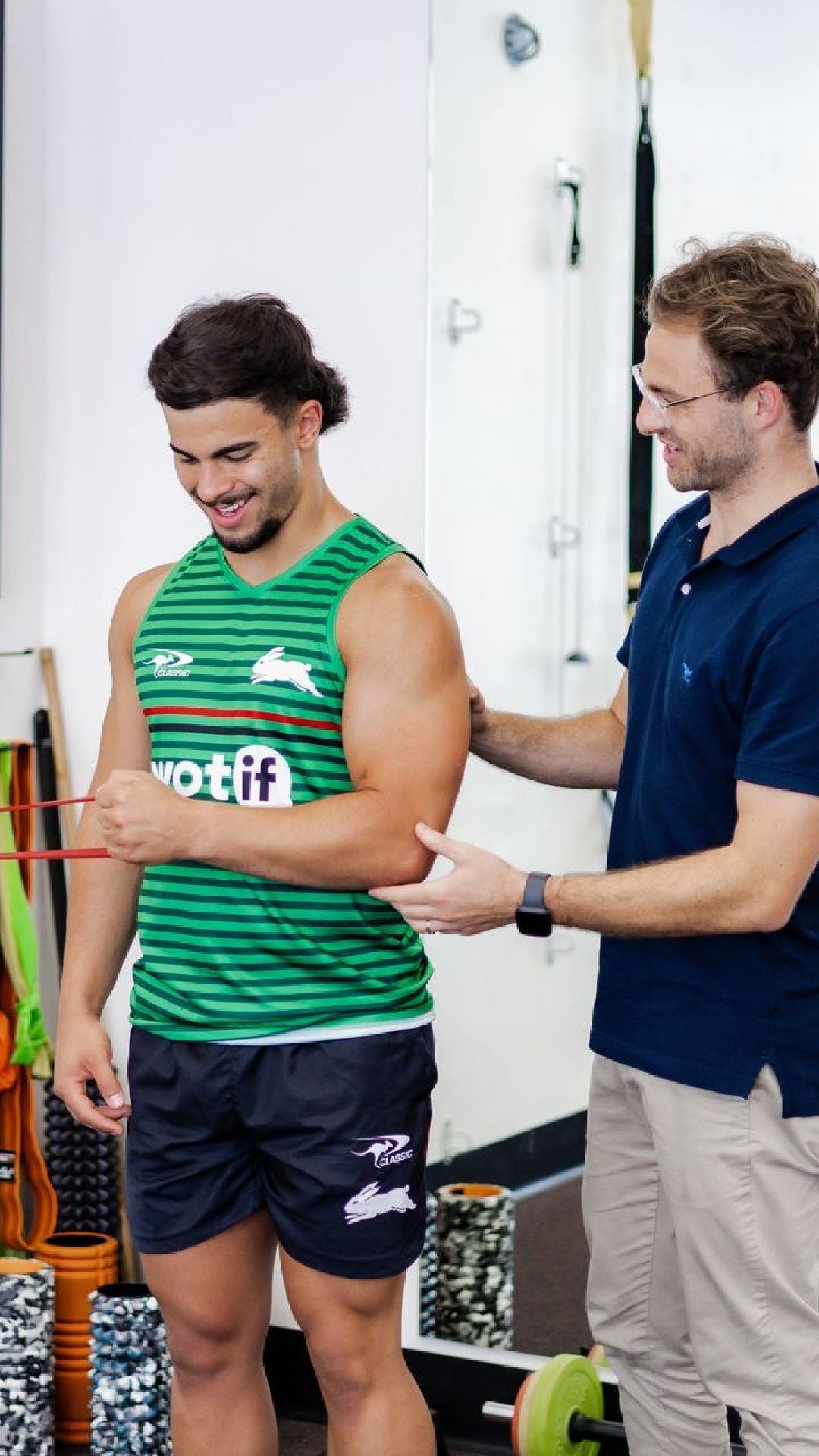 Man using resistance band during exercise, supervised by another man in a gym setting. Both are smiling.