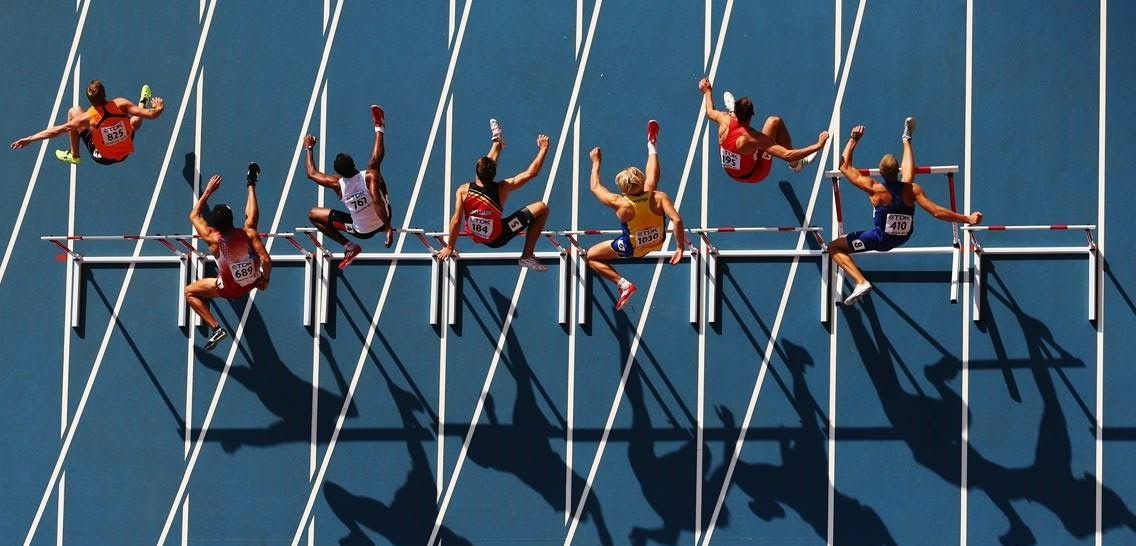 Athletes jumping hurdles in a track and field competition, blue track, sunny.