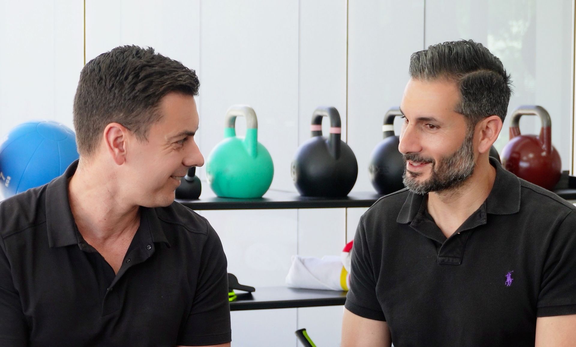 Two men in black shirts conversing in a gym, looking at each other. Kettlebells and a ball are in the background.