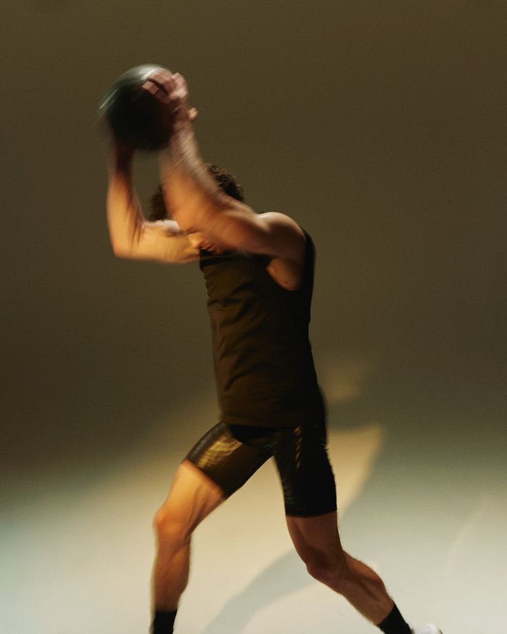 Athlete in black workout clothes leaping with a ball raised overhead against a plain studio background