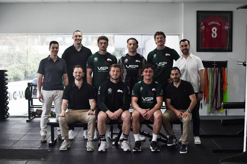 Group of men in a physical therapy clinic, some wearing sports team apparel, smiling for a photo.