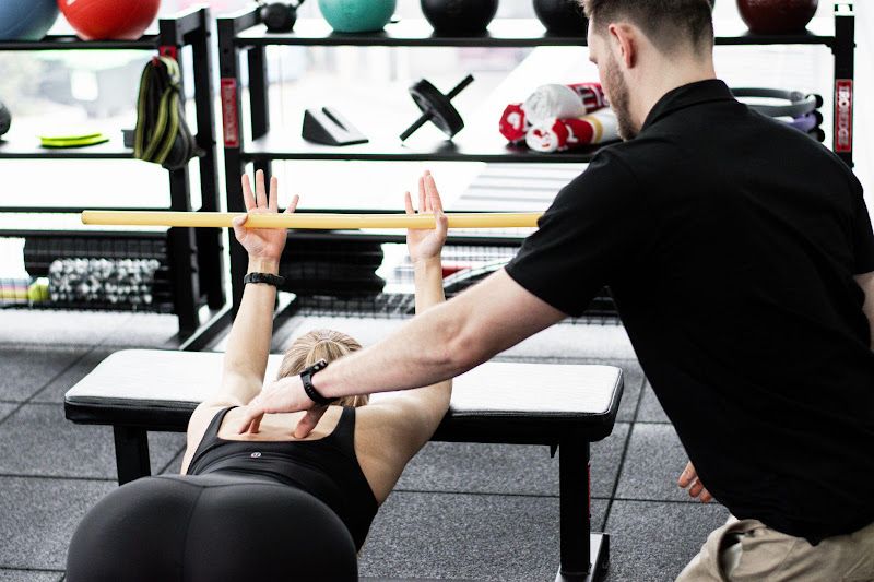 Woman doing a shoulder stretch with trainer, using a bar on a bench, in a gym.