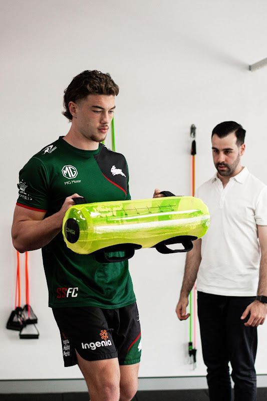 Man holding a yellow weight bag, being coached by another man in a gym setting.