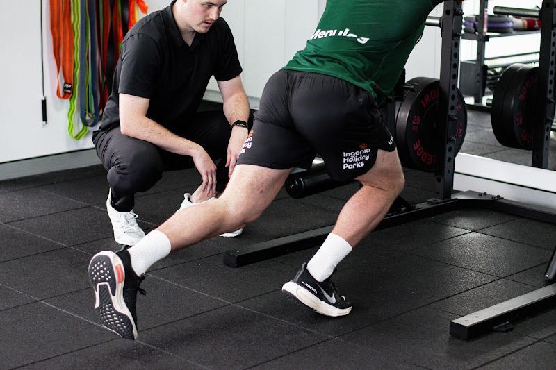 Man lunging with weight, supervised by a coach in a gym setting. Focus on form and exercise technique.