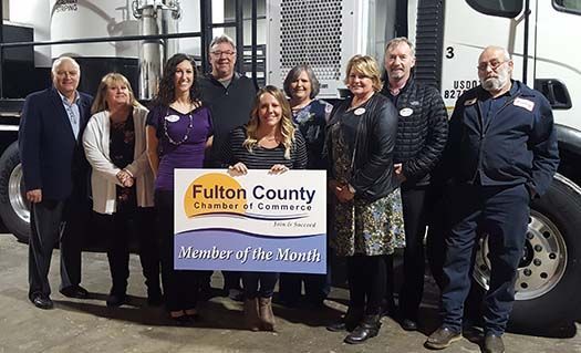 A group of people standing in front of a truck, holding a Fulton County Chamber of Commerce Member of the Month sign.
