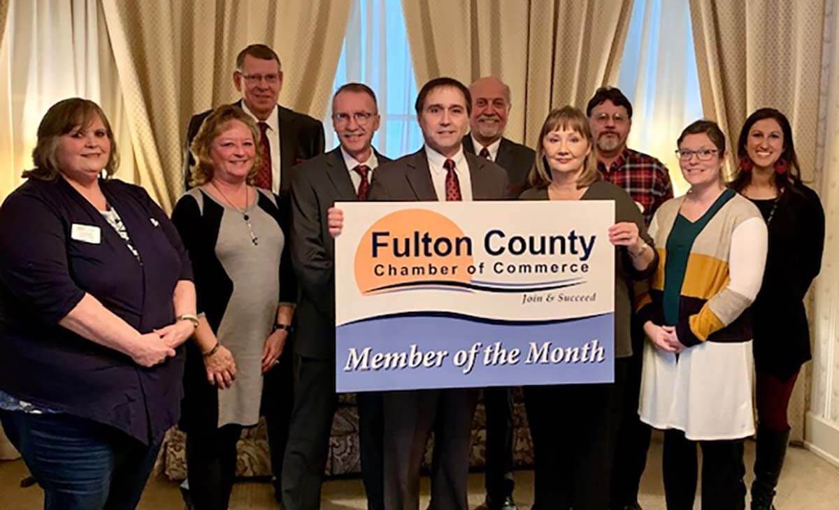 A group of people holding a Fulton County Chamber of Commerce Member of the Month sign in a room with curtained windows.