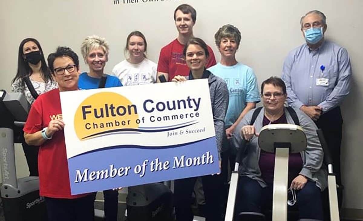 A group of nine people holding a Fulton County Chamber of Commerce Member of the Month sign in a gym-like setting.