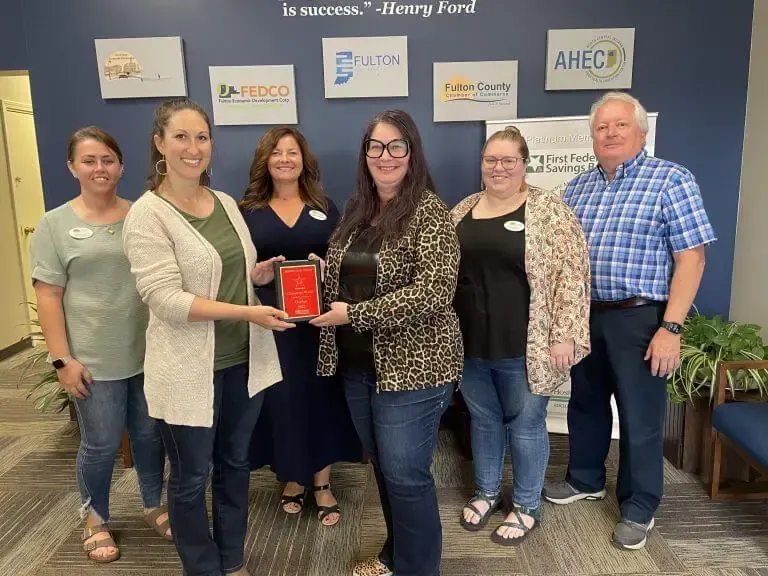 A group stands in an office in front of organizational logos; two women in the center hold a red plaque.