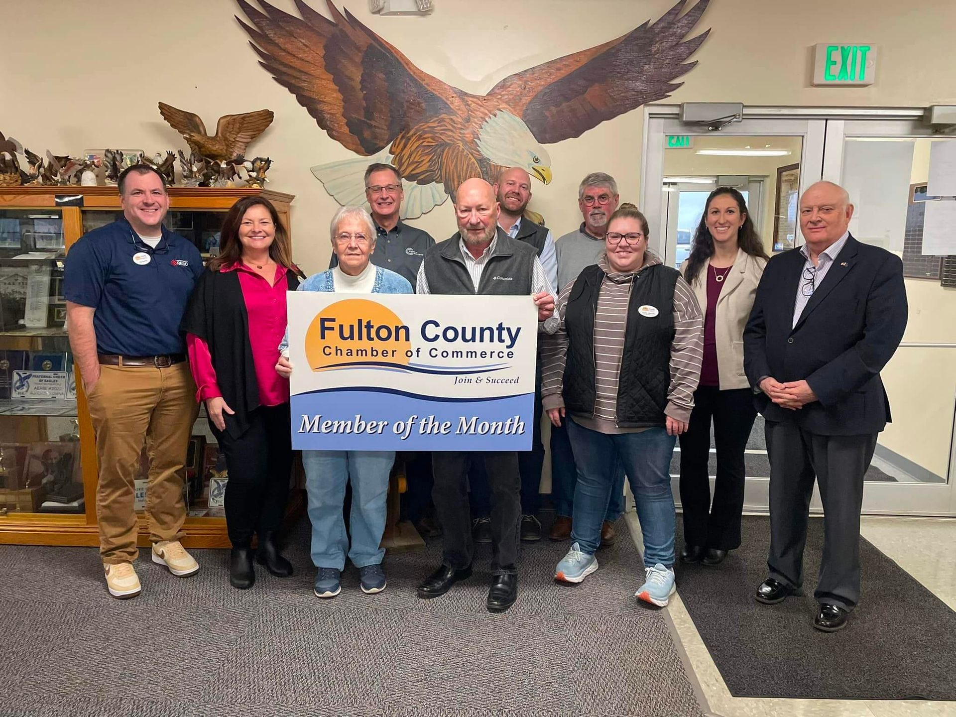 A group of people standing indoors holding a Fulton County Chamber of Commerce Member of the Month sign.