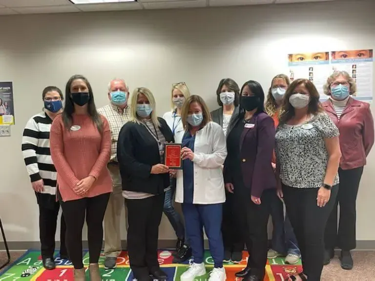 A group of people wearing face masks stands together in an office, with two people holding a red award plaque.