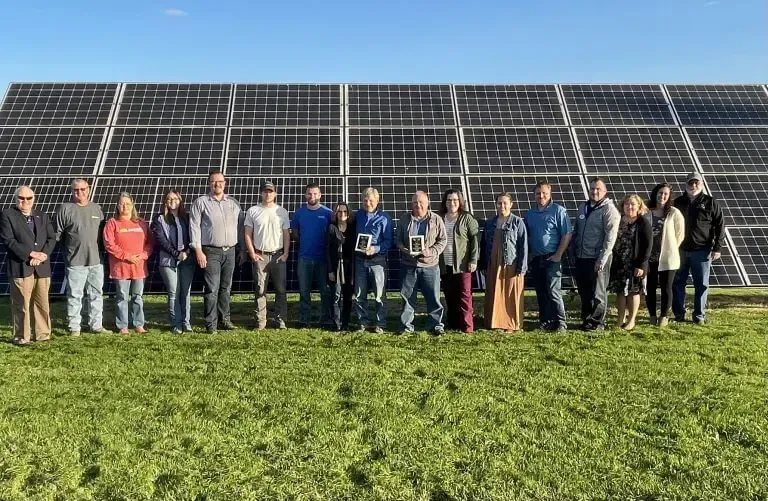 A group of people standing in a line in front of a large solar panel array on a grassy field under a clear blue sky.