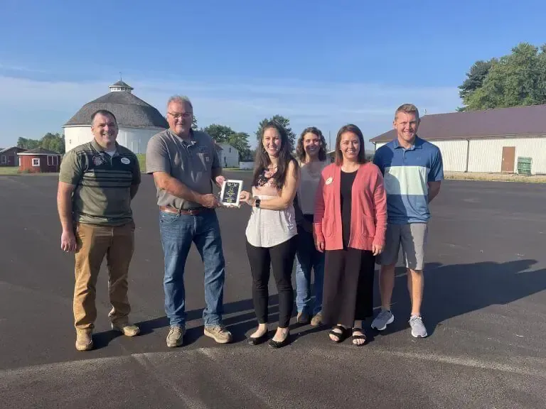 Six people stand in an outdoor parking lot with buildings in the background; a person in the center holds a small plaque.