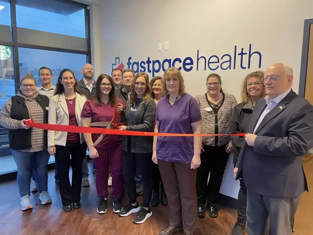 A group of people standing in a Fast Pace Health office, holding a red ribbon during a ribbon-cutting ceremony.