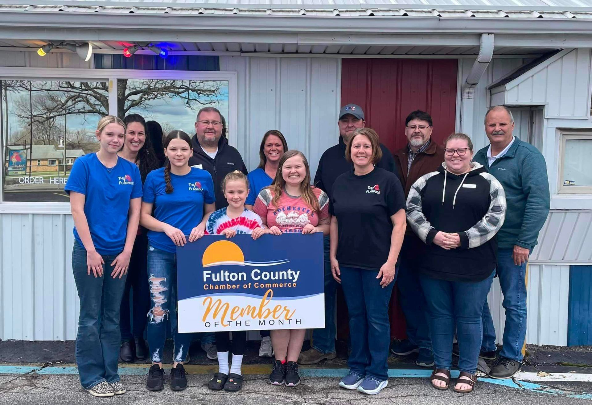A group of people standing in front of a building, holding a Fulton County Chamber of Commerce member sign.