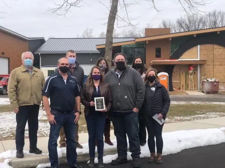 A group of people wearing face masks stands outdoors in front of a modern building, one person holding a plaque.
