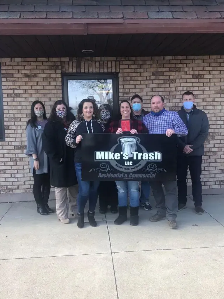 A group of eight people wearing masks stand in front of a brick building holding a Mike's Trash LLC sign.