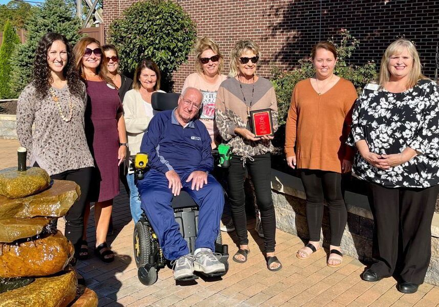 A group poses for a photo outdoors in front of a brick building and stone fountain, with one person holding an award plaque.
