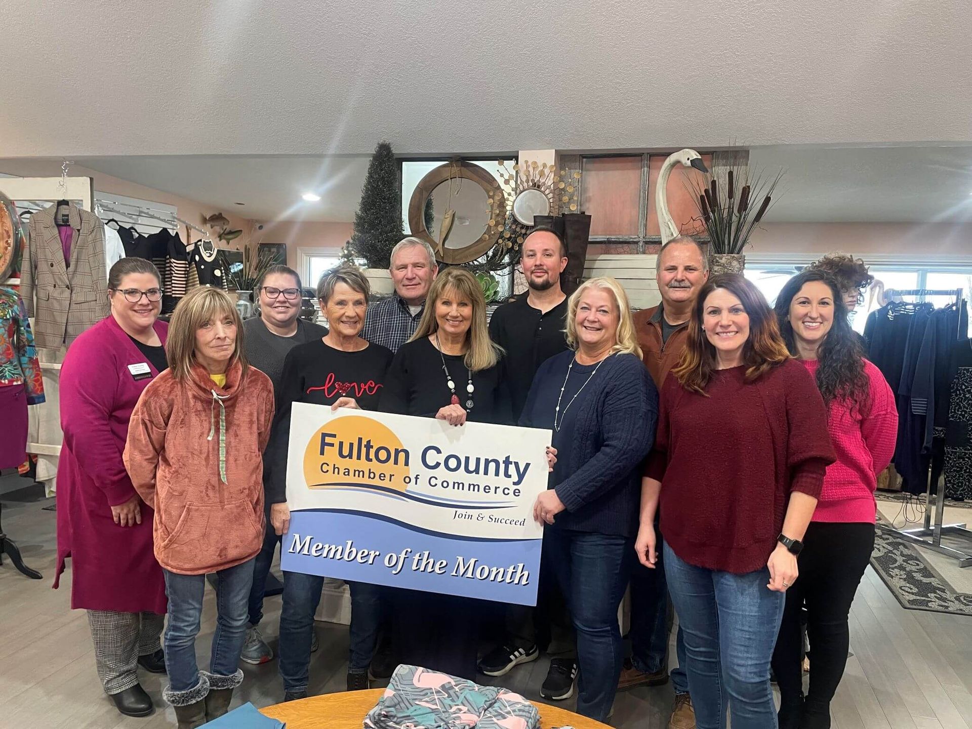 A group of twelve people standing in a shop, holding a Fulton County Chamber of Commerce Member of the Month sign.