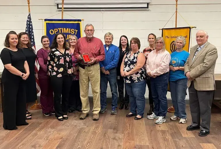 A group of people standing in a row in front of Optimist Club banners, with one person holding a small award plaque.