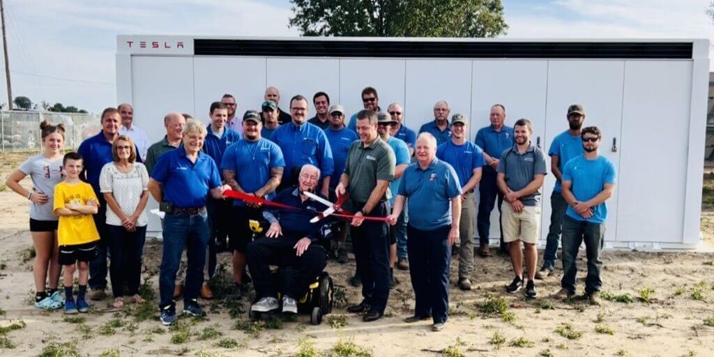 A group of people standing for a ribbon-cutting ceremony in front of a white Tesla energy storage unit outdoors.
