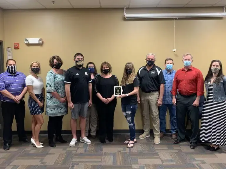 A group of eleven people stand in a line against a beige wall, with a central person holding a small award plaque.