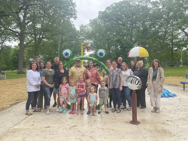 A group of people stands outdoors at a playground in front of a large, green, frog-shaped water feature.