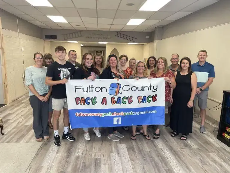 A group of people holding a banner for Fulton County Pack a Back Pack in a room with light-colored flooring.
