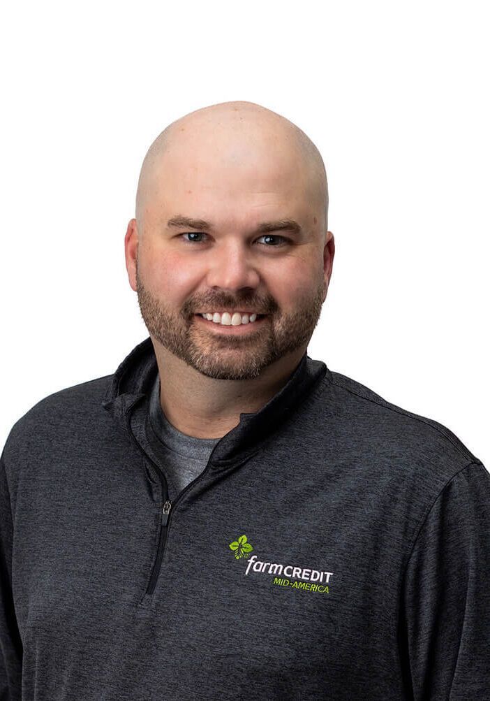 A smiling person with a shaved head and a beard wearing a dark grey, quarter-zip pullover against a white background.