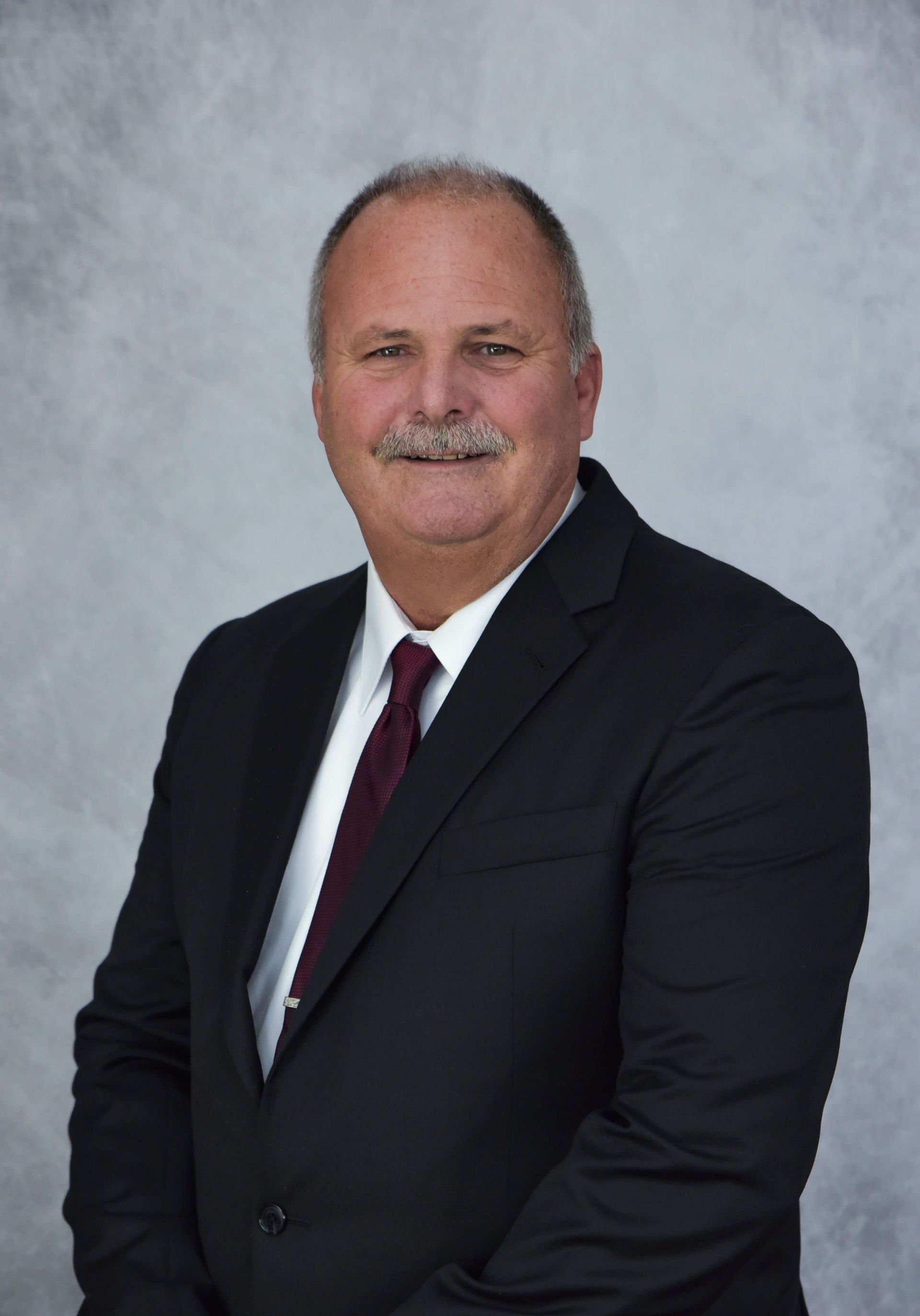 A man with a mustache in a dark suit, white shirt, and maroon tie posing against a light gray textured background.