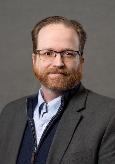 A professional headshot of a person with a beard and glasses, wearing a blazer, sweater, and light blue button-down shirt.
