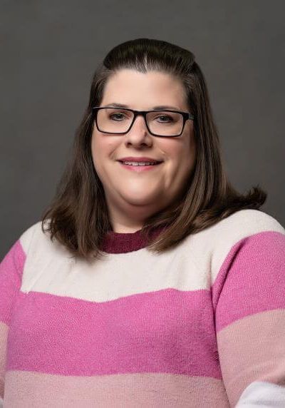 A person with shoulder-length brown hair and glasses wears a striped pink, white, and maroon sweater against a gray background.