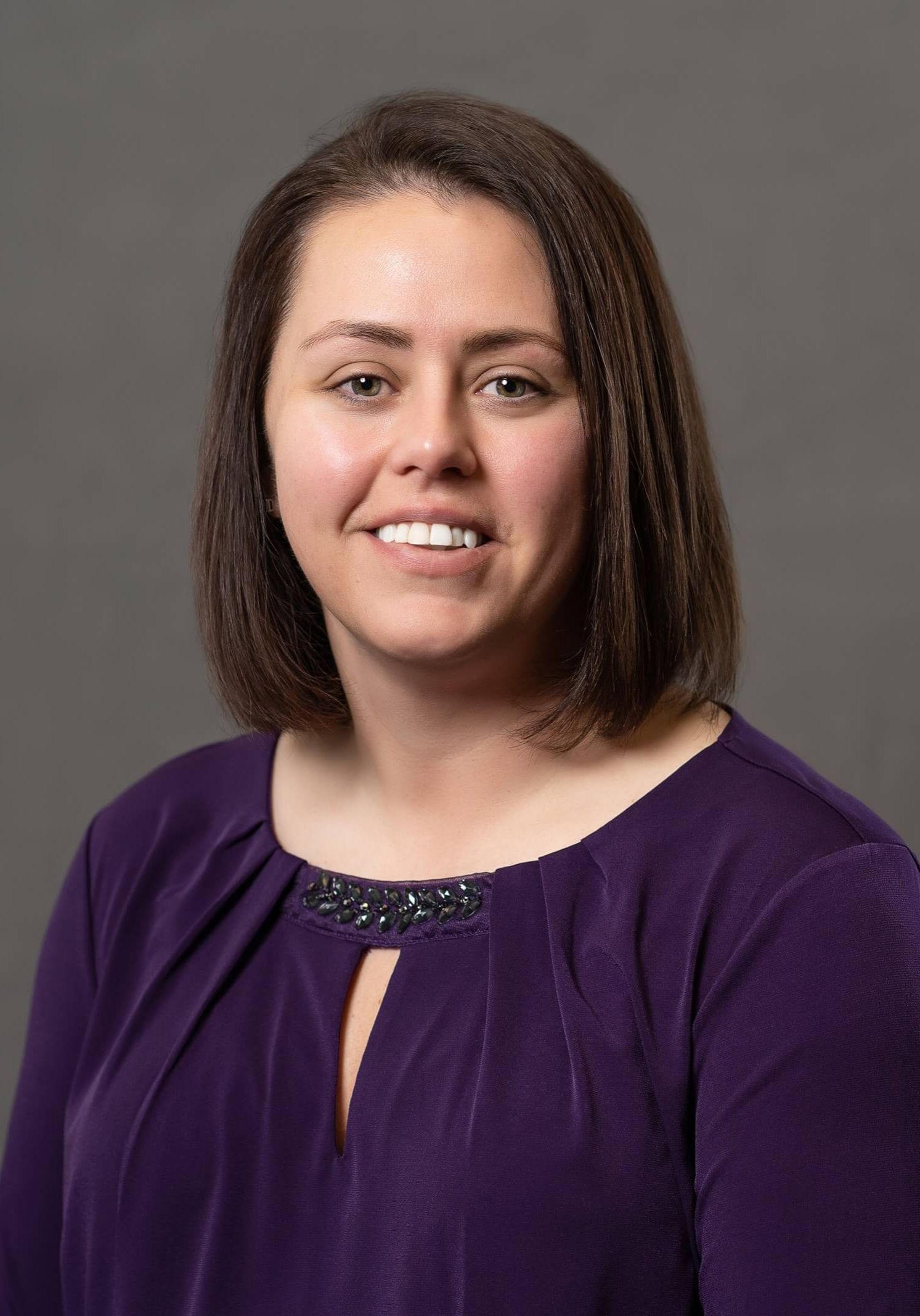 A smiling person wearing a dark purple top with a keyhole neckline, positioned against a plain grey background.