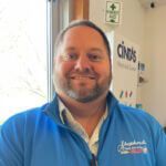 A smiling person with a beard wears a blue zip-up sweater in an office setting with a First Aid sign in the background.