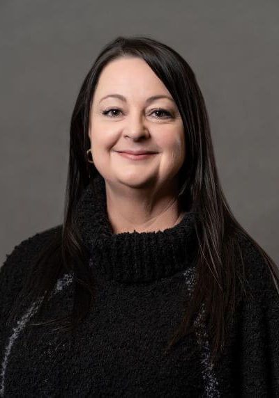 A person with long dark hair and a neutral expression, wearing a black textured cowl-neck sweater against a gray background.