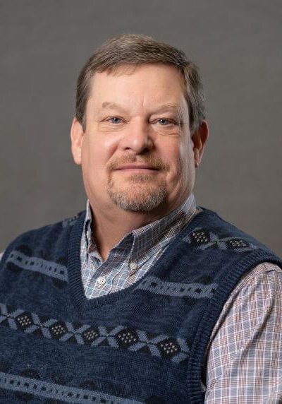 A person with brown hair and a goatee, wearing a patterned blue sweater vest over a plaid shirt against a gray background.