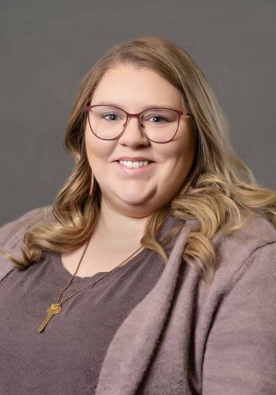 A person with long blonde hair and glasses wearing a mauve cardigan and a key pendant, smiling against a gray background.