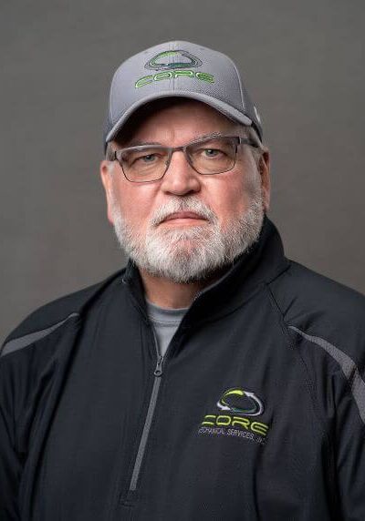 A person wearing a grey branded baseball cap, glasses, and a black quarter-zip jacket against a solid grey background.
