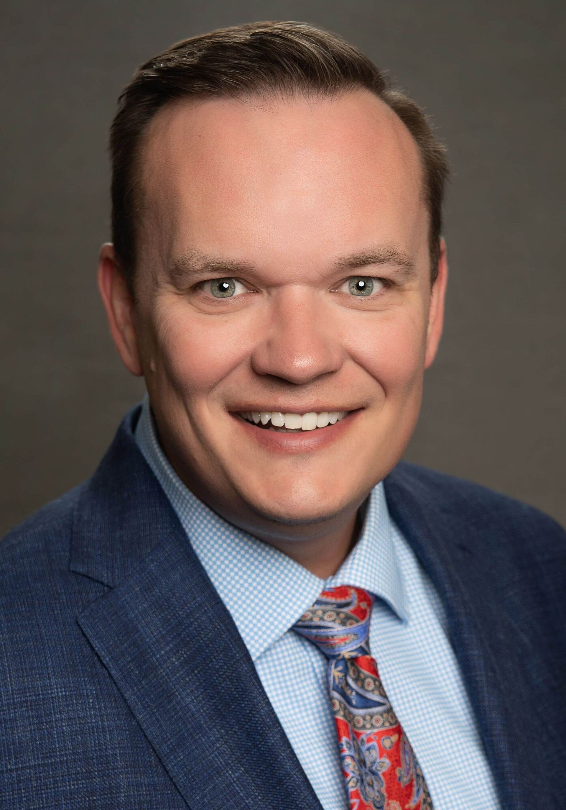 A smiling man wearing a blue suit, light blue patterned shirt, and a colorful paisley tie against a gray background.