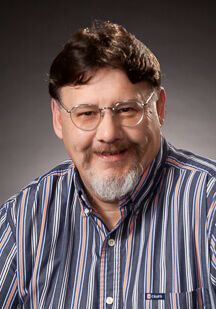 A person with a beard and glasses smiles, wearing a blue and white striped button-down shirt against a gray background.