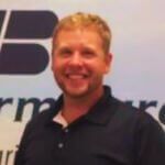 A person with short blond hair and a smiling expression, wearing a dark polo shirt in front of a branded background.