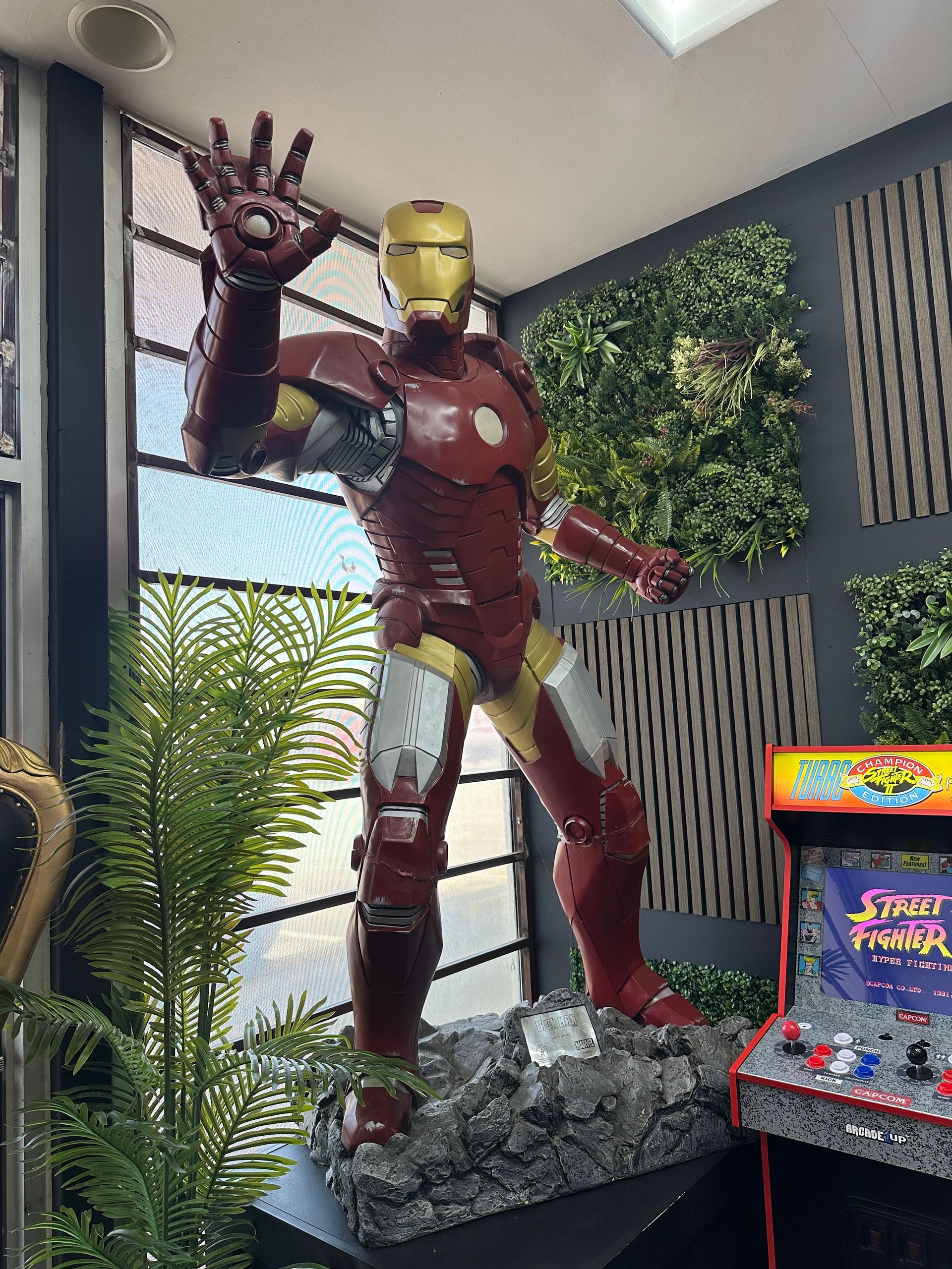 Iron Man statue in a display area, posed with raised hand beside an arcade cabinet and greenery.