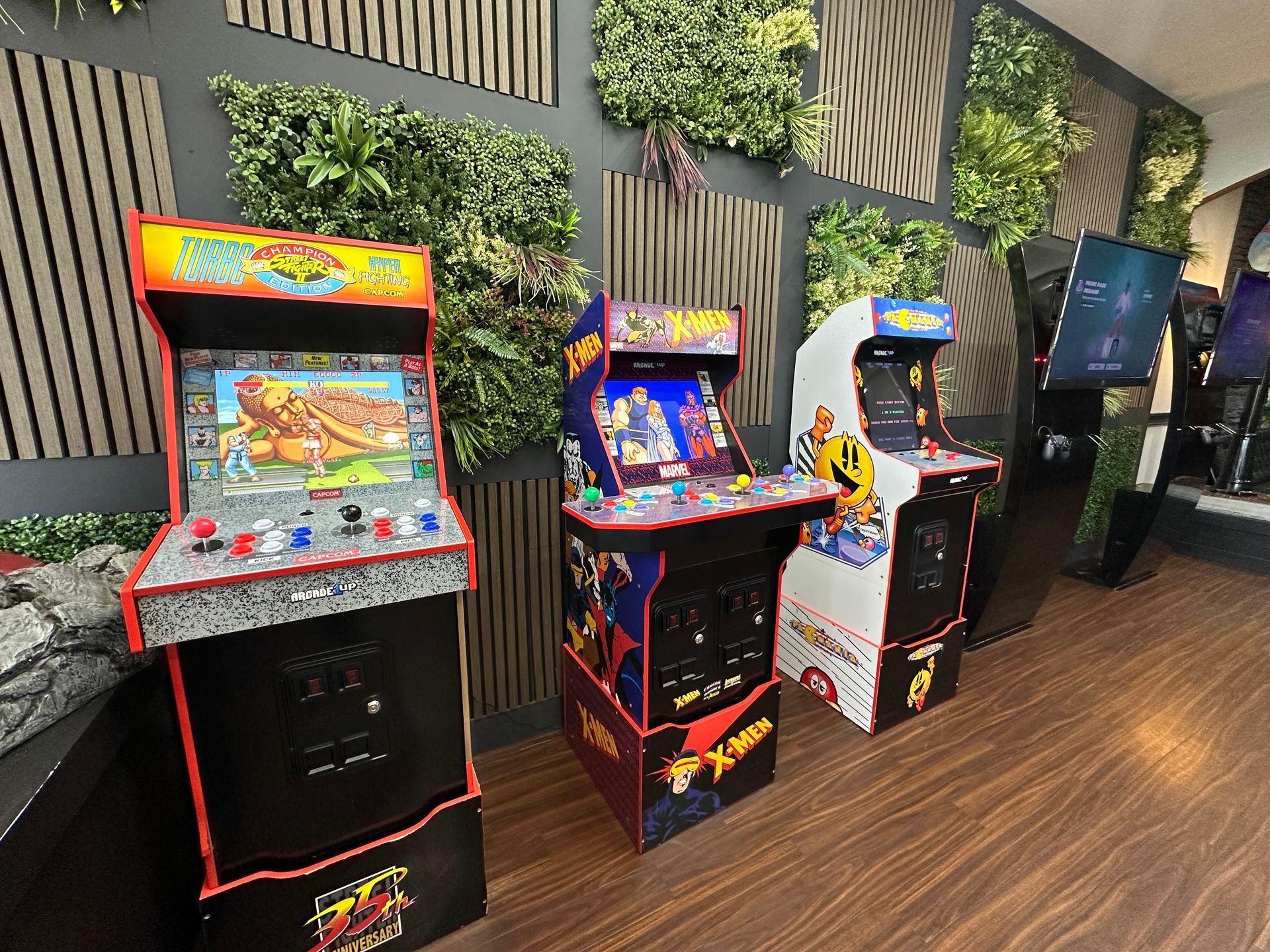 Arcade game cabinets lined up against a plant-covered wall in a modern lounge.