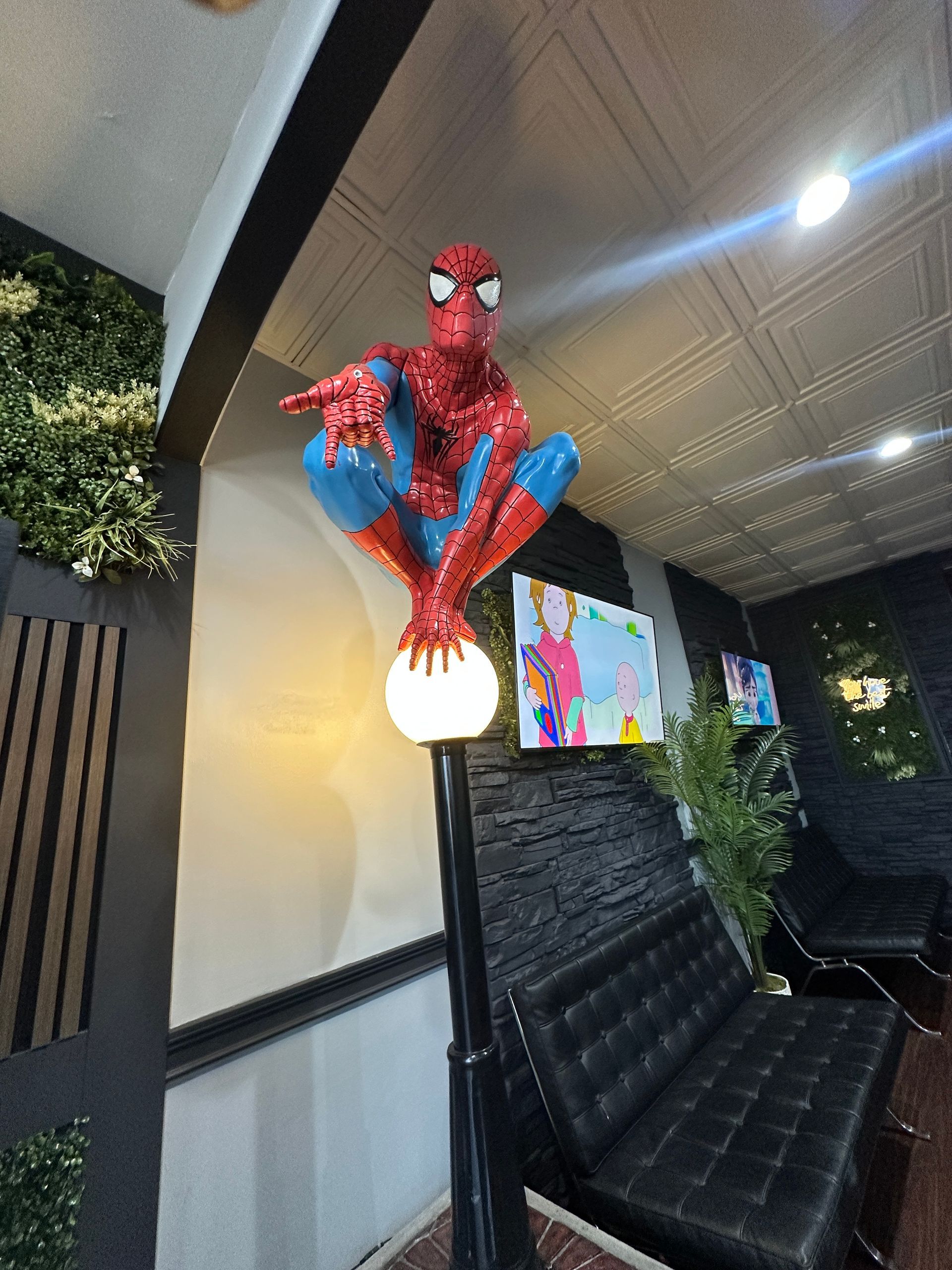 Spider-Man figure perched on a lamp in a modern indoor lounge with a wall-mounted TV.
