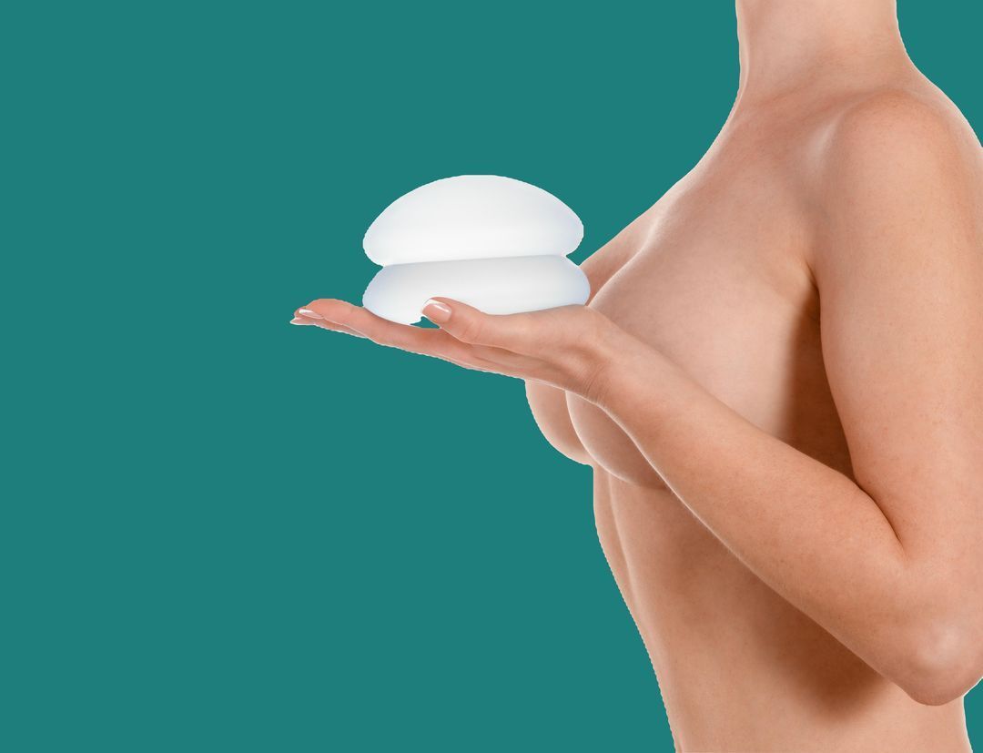 Woman holding two breast implants next to chest; turquoise background.