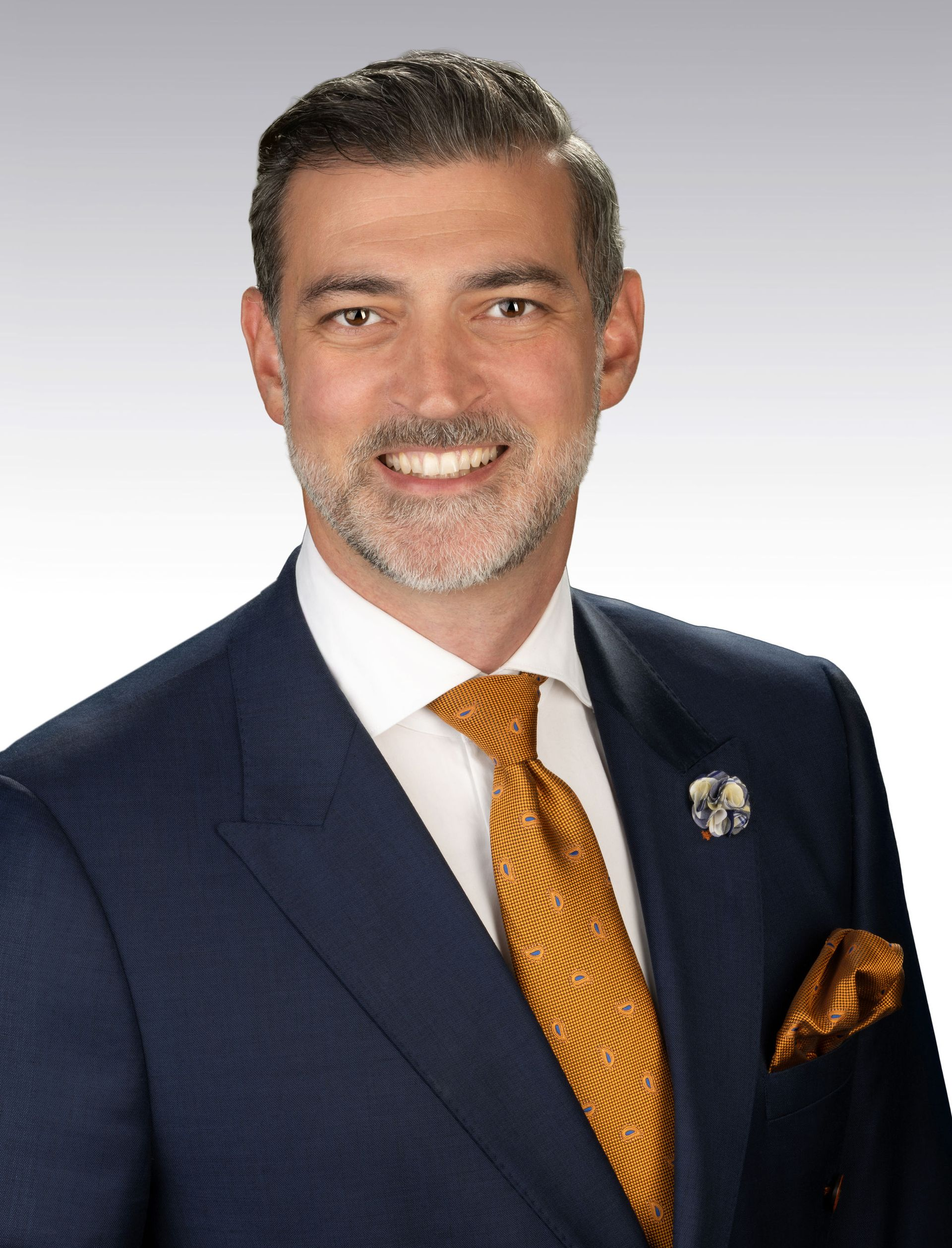 Man in blue suit, gold tie, smiling. Pocket square visible. Lapel pin.