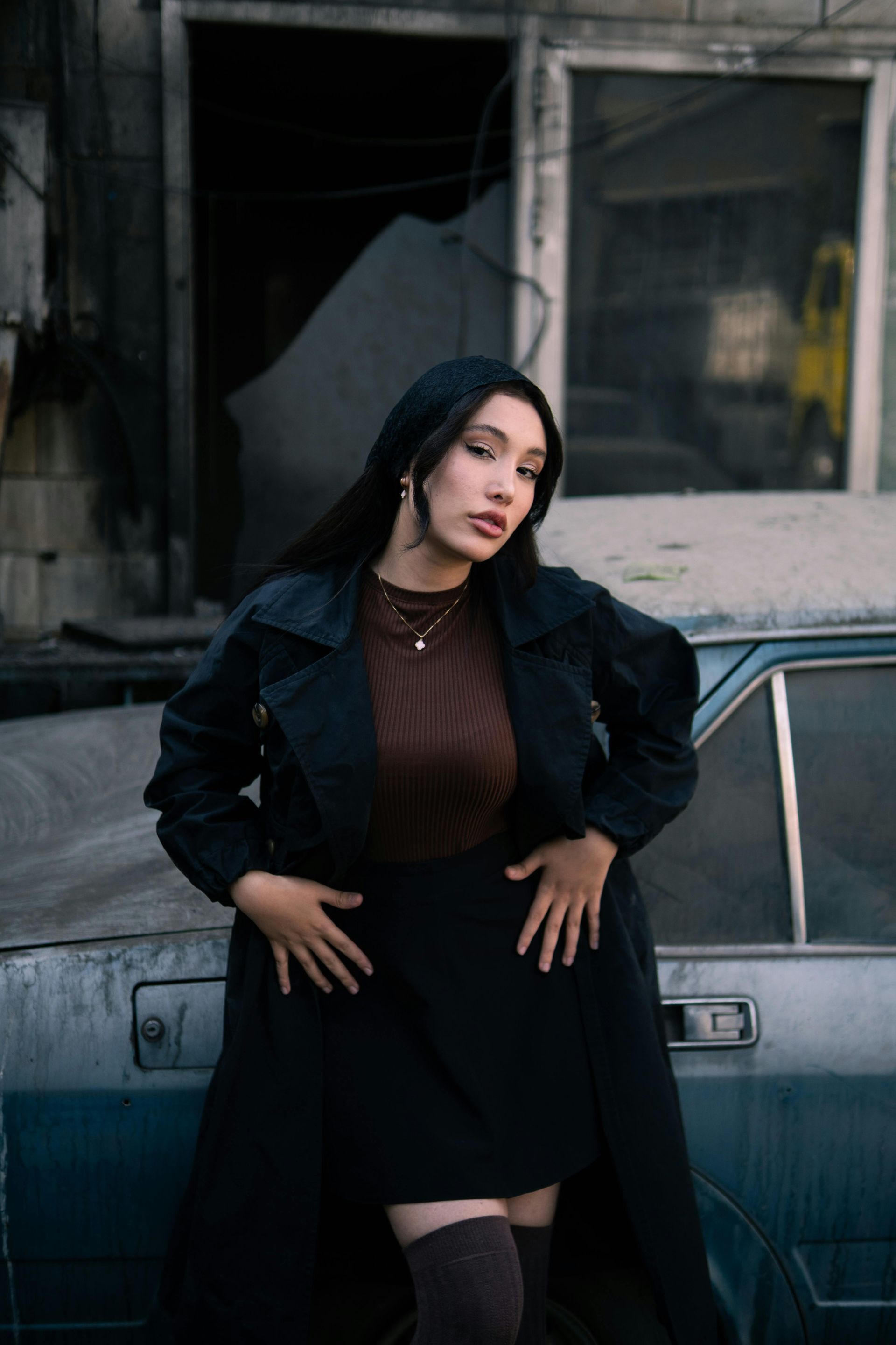 Woman in black coat and skirt, leaning on a weathered car; urban setting.