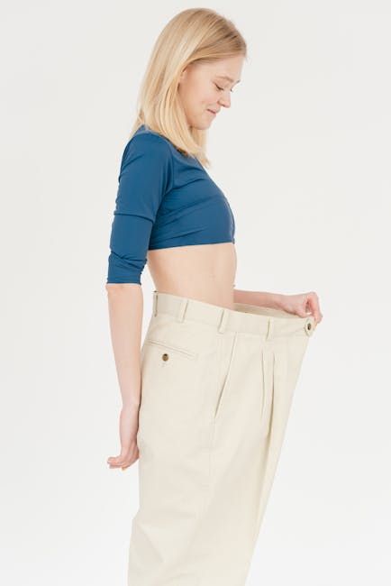 Woman wearing oversized pants, smiling, showing weight loss. Light-colored pants, blue top, white background.