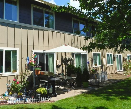 Backyard patio with table, umbrella, and grill, alongside a two-story building with tan siding and white-framed windows.